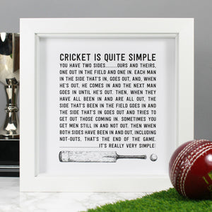 The 'Cricket is Quite Simple' Quote framed art print product photo shows the art print displayed in a white 8by8 box frame. The quote is black and white and incudes an ink illustration of a vintage cricket bat and cricket ball at the bottom of the art print. The framed print is displayed against a grey background with a trophy, and a red cricket ball and section of green astroturf in the foreground of the photograph.